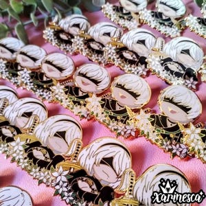 May include: A collection of white and gold enamel pins featuring a character with white hair and a black outfit. The pins are surrounded by white flowers and green leaves. The pins are arranged in rows on a pink surface. The text "xarinesca" is visible in the bottom right corner of the image.