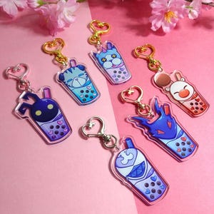 May include: Six colorful bubble tea charm keychains with different cartoon characters. Each keychain features a clear plastic charm with a cartoon character and a gold heart-shaped ring.