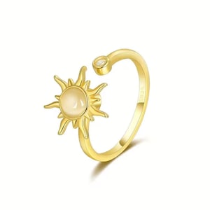 May include: A gold-colored ring featuring a sunburst design with a central, light-colored stone. The ring has an open design and a small, clear stone set into the band. The ring is likely made of gold-plated metal.