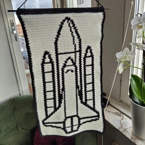 May include: A white crocheted wall hanging with a black outline of a space shuttle. The shuttle is made up of individual stitches and has a detailed design.