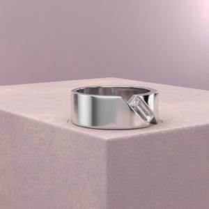 K&ouml;nnte beinhalten: Ein silberner Ring mit modernem Design. Der Ring hat einen rechteckigen, klaren Edelstein, der diagonal &uuml;ber ein breites Band gesetzt ist. Der Ring wird auf einer hellrosa Oberfl&auml;che vor einem sanften rosa Hintergrund pr&auml;sentiert.