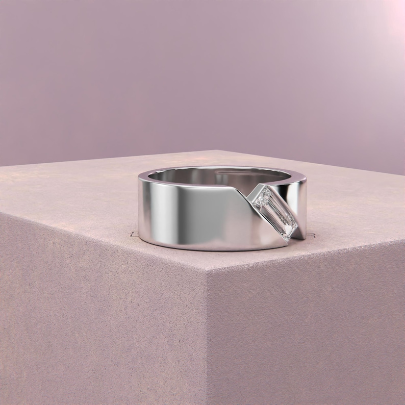K&ouml;nnte beinhalten: Ein silberner Ring mit modernem Design. Der Ring hat einen rechteckigen, klaren Edelstein, der diagonal &uuml;ber ein breites Band gesetzt ist. Der Ring wird auf einer hellrosa Oberfl&auml;che vor einem sanften rosa Hintergrund pr&auml;sentiert.