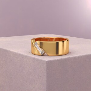 K&ouml;nnte beinhalten: Ein goldener Ring mit einem rechteckigen, klaren Edelstein, der diagonal &uuml;ber einem ausgeschnittenen Bereich eingesetzt ist. Der Ring ist dick und poliert und liegt auf einer hellgrauen Oberfl&auml;che vor einem weichen lila Hintergrund.