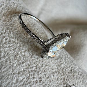 Puede incluir: Un anillo plateado con una gran piedra preciosa clara en forma de pera. La banda es delgada y lisa, mientras que el engaste alrededor de la piedra est&aacute; incrustado con piedras m&aacute;s peque&ntilde;as y brillantes. El anillo se encuentra sobre un fondo suave y texturizado.