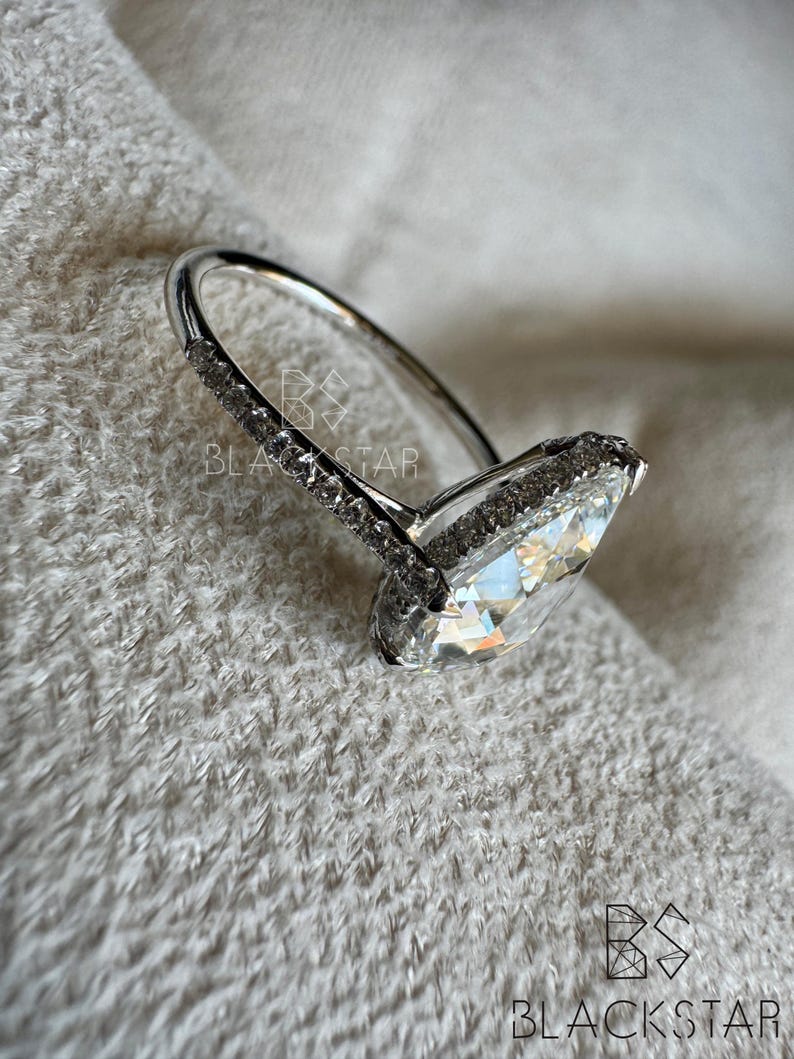Puede incluir: Un anillo plateado con una gran piedra preciosa clara en forma de pera. La banda es delgada y lisa, mientras que el engaste alrededor de la piedra est&aacute; incrustado con piedras m&aacute;s peque&ntilde;as y brillantes. El anillo se encuentra sobre un fondo suave y texturizado.