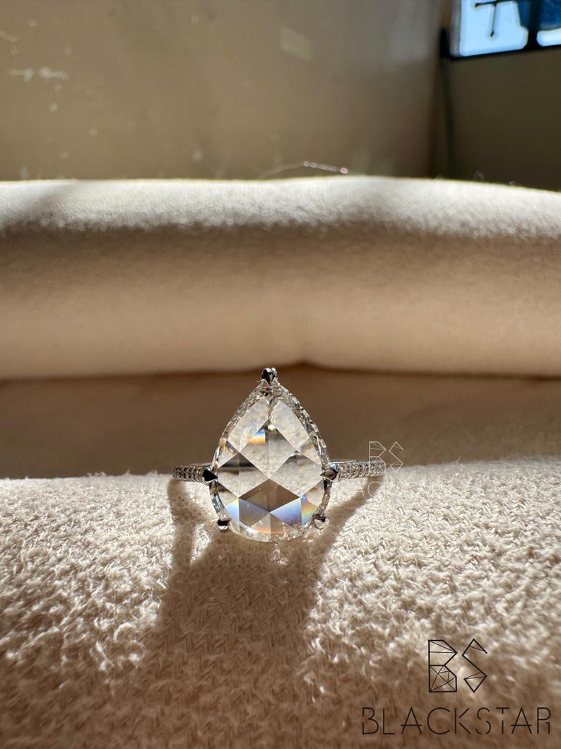 Front view of a pear shaped lab grown diamond engagement ring resting on a neutral jewelry cushion, displaying the brilliant pear cut center stone with a shimmering halo and pavé white gold band. The balanced proportions and exceptional sparkle highlight luxury craftsmanship and ethical sourcing. Perfect for buyers searching pear lab diamond engagement rings, eco friendly bridal jewelry, luxury diamond solitaires, and modern engagement ring styles.