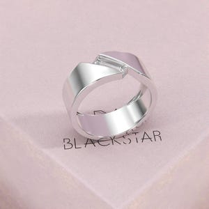 K&ouml;nnte beinhalten: Ein silberner Ring mit modernem Design. Der Ring hat eine geometrische Form mit einem kleinen, klaren Steinakzent. Das Wort "BLACKSTAR" ist im Hintergrund zu sehen. Der Ring wird auf einer rosa Oberfl&auml;che pr&auml;sentiert.