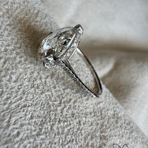 Puede incluir: Anillo de plata con un diamante en forma de pera, rodeado por un halo de diamantes m&aacute;s peque&ntilde;os. La banda tambi&eacute;n est&aacute; engastada con peque&ntilde;os diamantes. El anillo se presenta sobre un fondo blanco texturizado. El logotipo de Blackstar es visible.