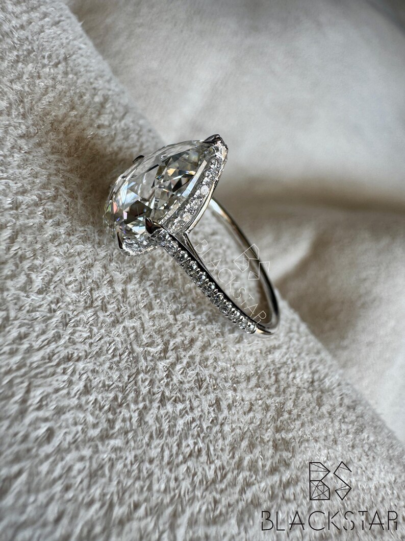 Puede incluir: Anillo de plata con un diamante en forma de pera, rodeado por un halo de diamantes m&aacute;s peque&ntilde;os. La banda tambi&eacute;n est&aacute; engastada con peque&ntilde;os diamantes. El anillo se presenta sobre un fondo blanco texturizado. El logotipo de Blackstar es visible.