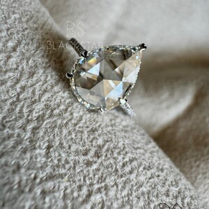 Puede incluir: Un anillo de plata con un diamante en forma de pera. La piedra clara y facetada es el punto focal, con piedras m&aacute;s peque&ntilde;as en la banda. El anillo descansa sobre una superficie suave y texturizada. La marca "BLACKSTAR" es visible.
