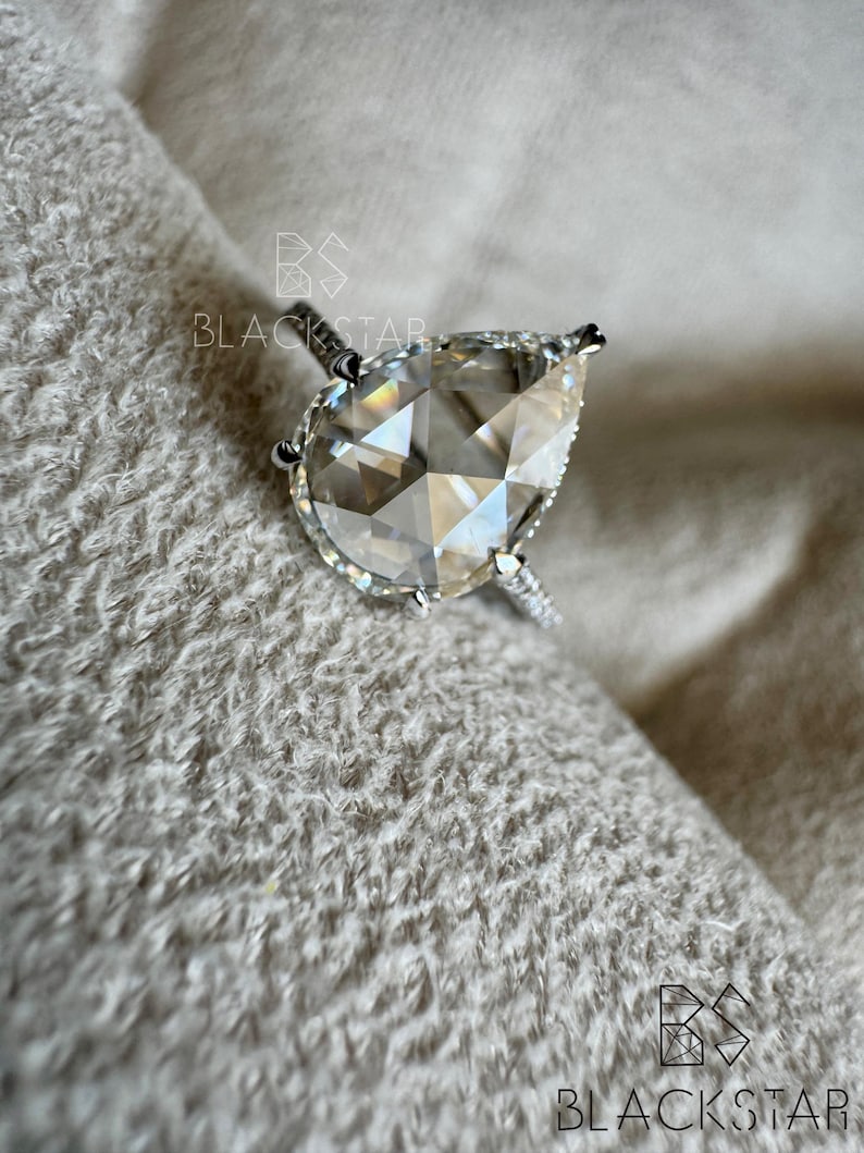 Puede incluir: Un anillo de plata con un diamante en forma de pera. La piedra clara y facetada es el punto focal, con piedras m&aacute;s peque&ntilde;as en la banda. El anillo descansa sobre una superficie suave y texturizada. La marca "BLACKSTAR" es visible.