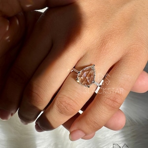 Puede incluir: Anillo de plata con una gema en forma de pera. El anillo tiene un halo de piedras m&aacute;s peque&ntilde;as alrededor de la gema principal. El anillo est&aacute; en una mano, con el logotipo de Blackstar en el fondo.