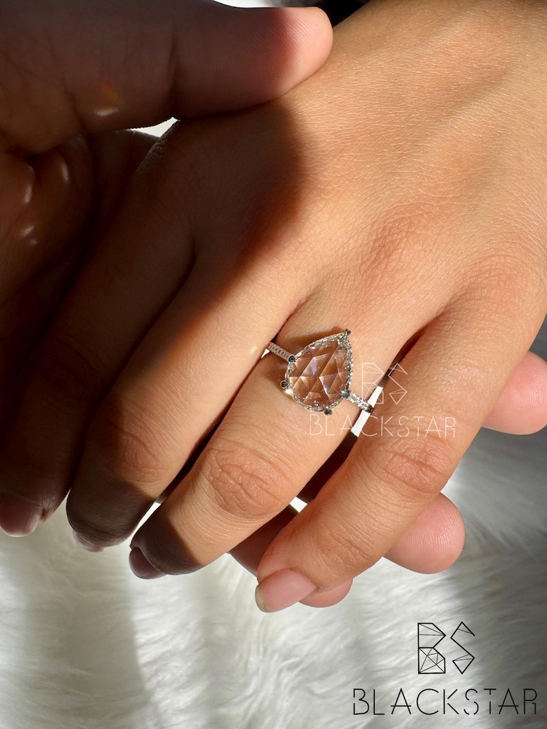 Puede incluir: Anillo de plata con una gema en forma de pera. El anillo tiene un halo de piedras m&aacute;s peque&ntilde;as alrededor de la gema principal. El anillo est&aacute; en una mano, con el logotipo de Blackstar en el fondo.