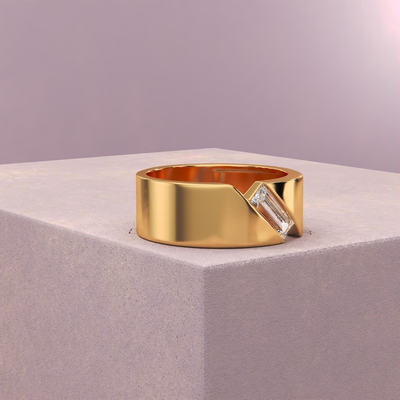 K&ouml;nnte beinhalten: Ein goldener Ring mit einem rechteckigen Diamanten, der schr&auml;g eingesetzt ist. Der Ring hat ein breites Band und ein modernes Design. Der Ring wird auf einer hellgrauen Oberfl&auml;che vor einem weichen lila Hintergrund pr&auml;sentiert.