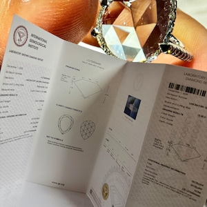 Close-up image of an IGI certified pear shaped lab grown diamond engagement ring displayed alongside its official International Gemological Institute laboratory grown diamond report. The pear cut diamond features exceptional brilliance, halo detailing, and pavé white gold band craftsmanship. This certified lab grown diamond ring highlights authenticity, ethical sourcing, and premium quality, ideal for buyers searching IGI certified lab diamond rings, pear shape engagement rings, fine jewelry