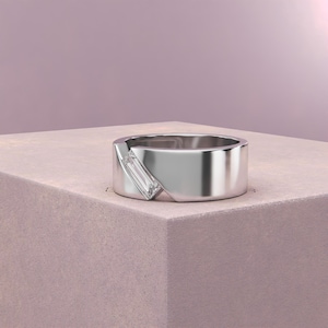 K&ouml;nnte beinhalten: Ein silberner Ring mit einem rechteckigen, klaren Edelstein, der diagonal &uuml;ber das Band gesetzt ist. Der Ring hat ein modernes, minimalistisches Design und liegt auf einer rosa Oberfl&auml;che.
