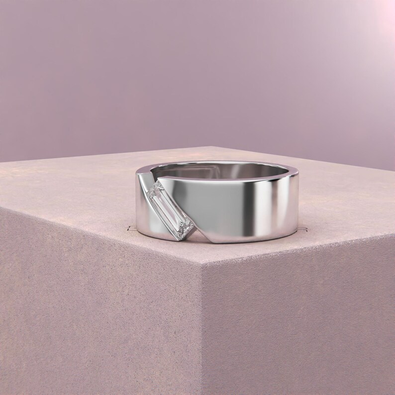 K&ouml;nnte beinhalten: Ein silberner Ring mit einem rechteckigen, klaren Edelstein, der diagonal &uuml;ber das Band gesetzt ist. Der Ring hat ein modernes, minimalistisches Design und liegt auf einer rosa Oberfl&auml;che.