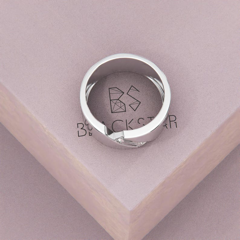 K&ouml;nnte beinhalten: Ein silberner Ring mit einem einzigartigen, ineinandergreifenden Design. Der Ring liegt auf einer hellvioletten Oberfl&auml;che mit dem Text "BLACKSTAR". Die polierte Oberfl&auml;che des Rings reflektiert das Licht und unterstreicht seine moderne &Auml;sthetik.