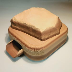 May include: A light brown wooden base with a textured surface and a small opening on the side. A light brown rock sits on top of the base.