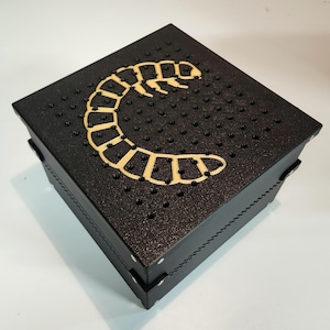 May include: A black square box with a gold-colored centipede design on the top. The box has a textured surface and a rope-like trim around the edges.