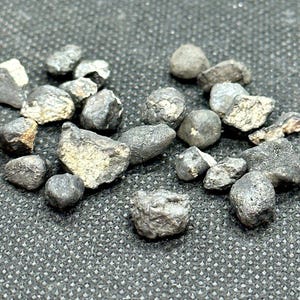 Pode incluir: Uma coleção de pequenos fragmentos de meteorito metálico cinza escuro. Os fragmentos têm uma forma irregular e uma superfície áspera e texturizada.