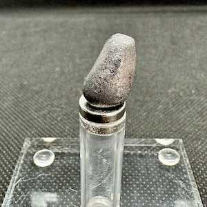 Könnte beinhalten: Ein kleiner, dunkelgrauer, unregelmäßig geformter Stein liegt auf einem durchsichtigen Glasfläschchen mit einem silbernen Deckel. Das Fläschchen steht auf einer durchsichtigen Glasfläche.