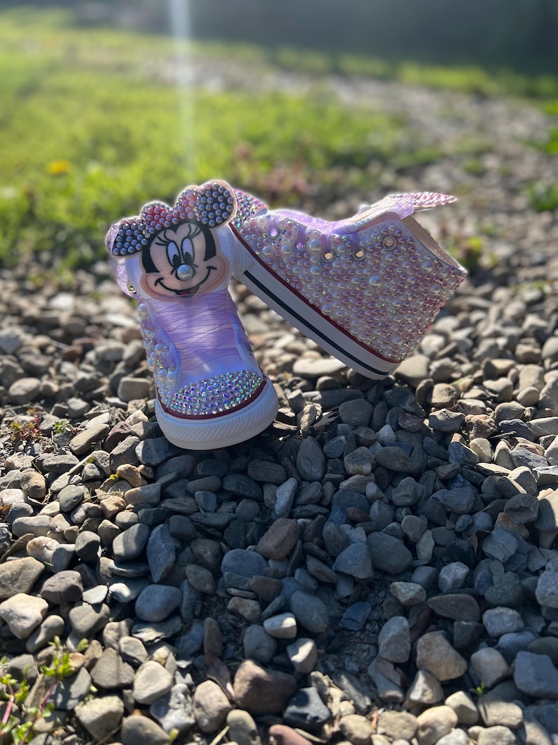 May include: A pair of custom-designed sneakers featuring a Minnie Mouse graphic on the tongue. The shoes are embellished with iridescent rhinestones, with a white rubber sole and a red stripe.