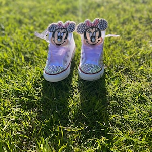 May include: A pair of white high-top sneakers with Minnie Mouse faces on the tongues. The shoes have pink laces and are embellished with rhinestones around the toe and on the Minnie Mouse bows. The shoes are sitting on green grass.