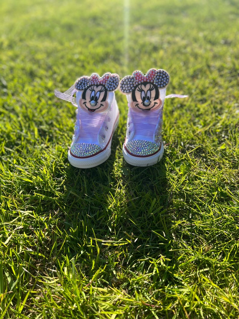 May include: A pair of white high-top sneakers with Minnie Mouse faces on the tongues. The shoes have pink laces and are embellished with rhinestones around the toe and on the Minnie Mouse bows. The shoes are sitting on green grass.