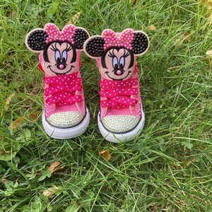 Puede incluir: Un par de zapatillas de lona rosa con adornos de Minnie Mouse. Cada zapato presenta una cara de Minnie Mouse en la leng&uuml;eta, cordones rosas con lunares y una puntera enjoyada. Las zapatillas est&aacute;n sobre hierba verde.