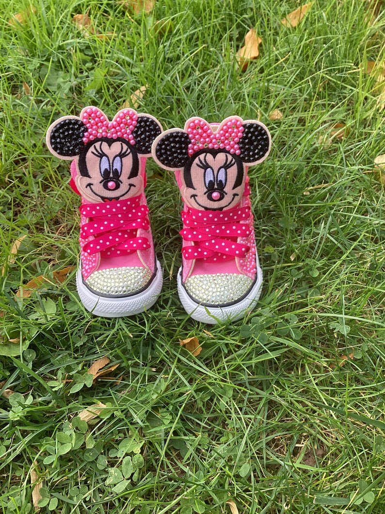 Puede incluir: Un par de zapatillas de lona rosa con adornos de Minnie Mouse. Cada zapato presenta una cara de Minnie Mouse en la leng&uuml;eta, cordones rosas con lunares y una puntera enjoyada. Las zapatillas est&aacute;n sobre hierba verde.