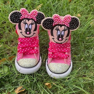 Puede incluir: Par de zapatillas rosas para ni&ntilde;os peque&ntilde;os con caras de Minnie Mouse en las leng&uuml;etas. Los zapatos tienen cordones de lunares, punteras con pedrer&iacute;a y suelas blancas. Las caras de Minnie Mouse tienen orejas negras y lazos rosas.