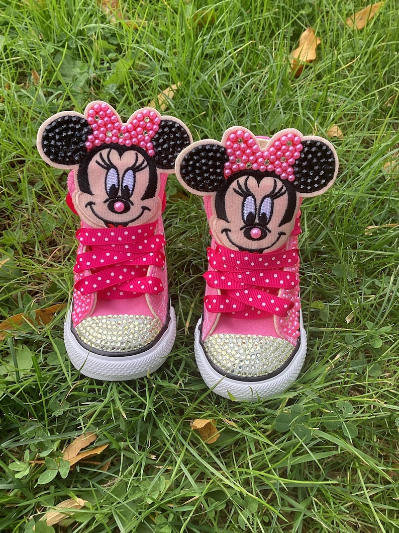 Puede incluir: Par de zapatillas rosas para ni&ntilde;os peque&ntilde;os con caras de Minnie Mouse en las leng&uuml;etas. Los zapatos tienen cordones de lunares, punteras con pedrer&iacute;a y suelas blancas. Las caras de Minnie Mouse tienen orejas negras y lazos rosas.