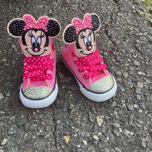 Puede incluir: Un par de zapatillas altas rosas con un dise&ntilde;o de Minnie Mouse. Los zapatos tienen suela blanca, punteras con pedrer&iacute;a y cordones rosas con lunares. La cara y el lazo de Minnie est&aacute;n en la leng&uuml;eta.