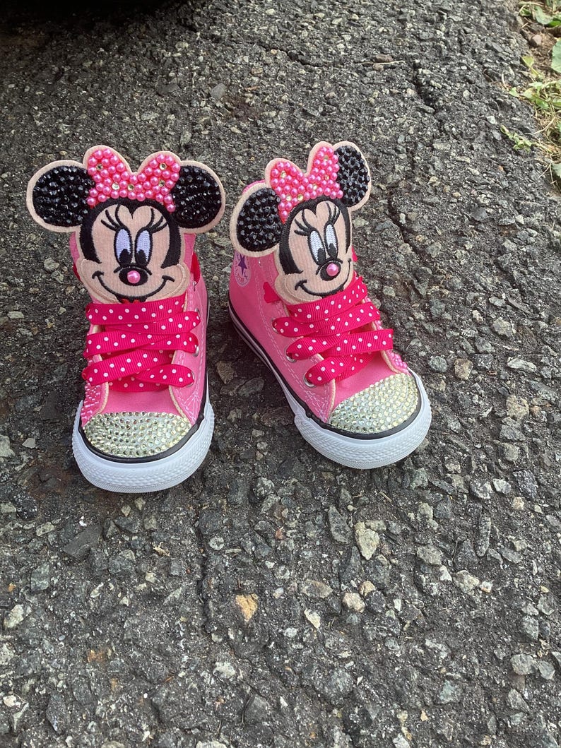 Puede incluir: Un par de zapatillas altas rosas con un dise&ntilde;o de Minnie Mouse. Los zapatos tienen suela blanca, punteras con pedrer&iacute;a y cordones rosas con lunares. La cara y el lazo de Minnie est&aacute;n en la leng&uuml;eta.
