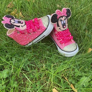 Puede incluir: Un par de zapatillas rosas para ni&ntilde;os peque&ntilde;os con suelas blancas y punteras doradas. Las zapatillas est&aacute;n adornadas con pedrer&iacute;a y presentan apliques del personaje de Minnie Mouse en las leng&uuml;etas. Los cordones rosas de lunares completan el dise&ntilde;o.