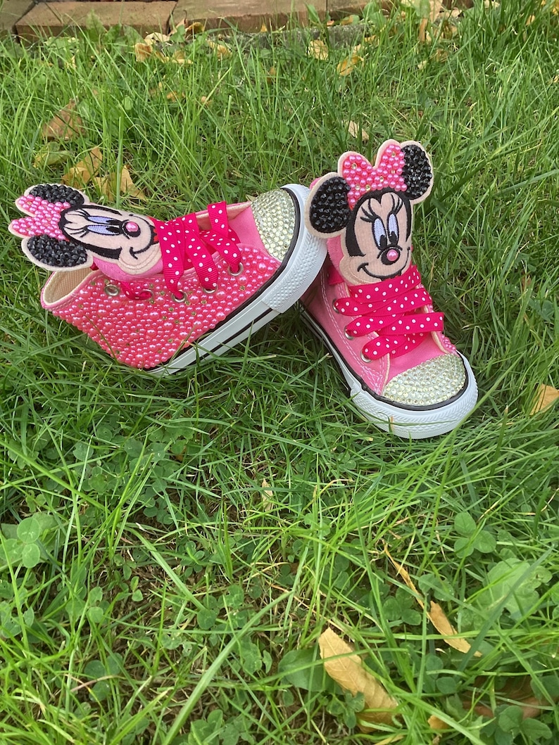 Puede incluir: Un par de zapatillas rosas para ni&ntilde;os peque&ntilde;os con suelas blancas y punteras doradas. Las zapatillas est&aacute;n adornadas con pedrer&iacute;a y presentan apliques del personaje de Minnie Mouse en las leng&uuml;etas. Los cordones rosas de lunares completan el dise&ntilde;o.