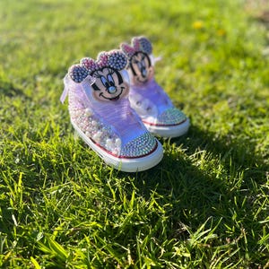 May include: A pair of custom-designed high-top sneakers featuring a Minnie Mouse theme. The shoes are primarily white and lavender, adorned with rhinestones and pearls. The design includes a Minnie Mouse face on the tongue and a red trim around the sole.