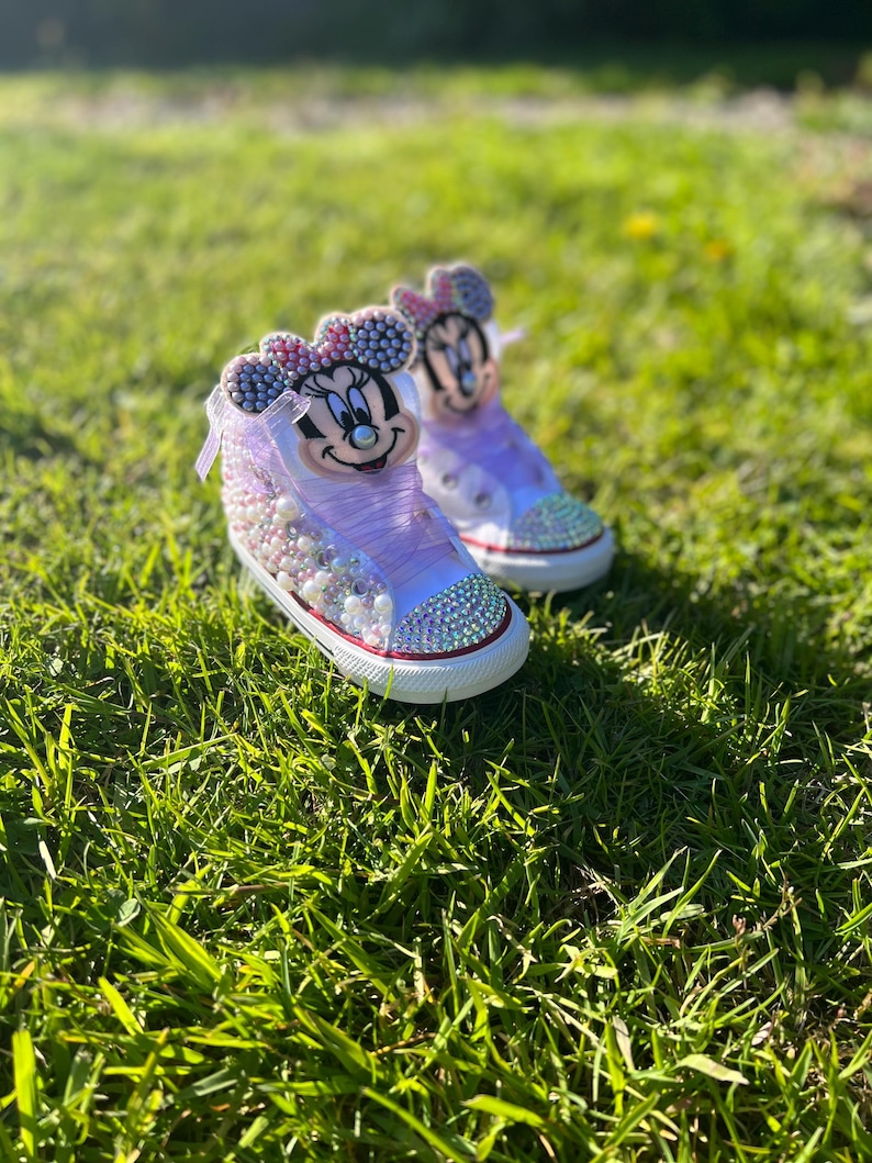 May include: A pair of custom-designed high-top sneakers featuring a Minnie Mouse theme. The shoes are primarily white and lavender, adorned with rhinestones and pearls. The design includes a Minnie Mouse face on the tongue and a red trim around the sole.