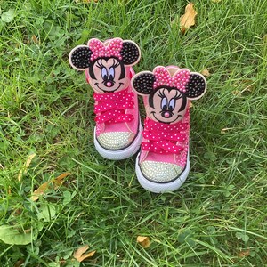 Puede incluir: Un par de zapatillas rosas para ni&ntilde;os peque&ntilde;os con caras de Minnie Mouse en las leng&uuml;etas. Los zapatos tienen suelas blancas, punteras cubiertas de pedrer&iacute;a y cordones rosas con lunares. Los zapatos est&aacute;n sobre hierba verde.