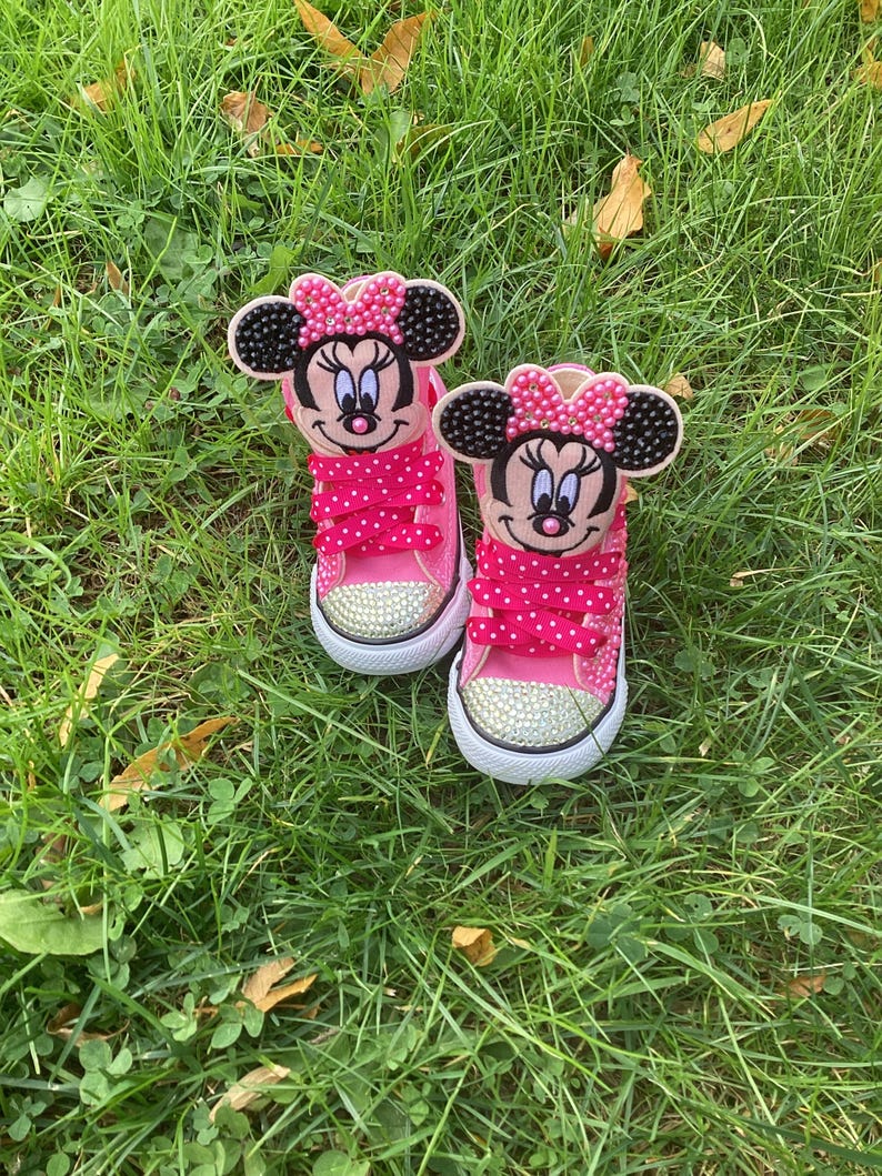 Puede incluir: Un par de zapatillas rosas para ni&ntilde;os peque&ntilde;os con caras de Minnie Mouse en las leng&uuml;etas. Los zapatos tienen suelas blancas, punteras cubiertas de pedrer&iacute;a y cordones rosas con lunares. Los zapatos est&aacute;n sobre hierba verde.