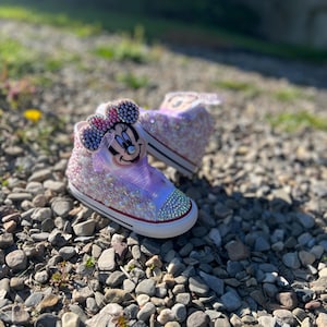 May include: A pair of high-top sneakers, embellished with sparkling rhinestones and a Minnie Mouse design. The shoes feature a white rubber sole, a pink and purple color scheme, and a bow on the top. The shoes are designed for children.