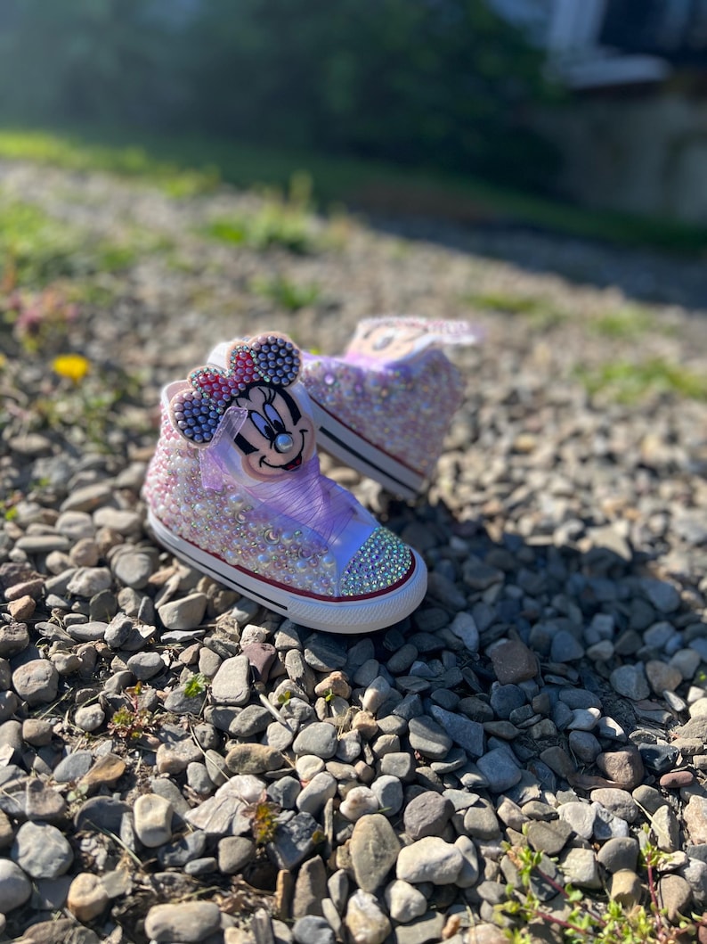 May include: A pair of high-top sneakers, embellished with sparkling rhinestones and a Minnie Mouse design. The shoes feature a white rubber sole, a pink and purple color scheme, and a bow on the top. The shoes are designed for children.