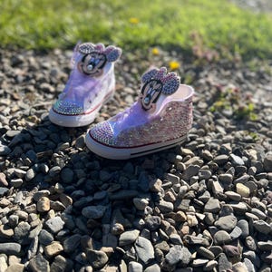 May include: A pair of custom high-top sneakers with a Minnie Mouse theme. The shoes are decorated with pink and silver glitter, rhinestones, and a Minnie Mouse face on the tongue. The shoes are sitting on a bed of small rocks.