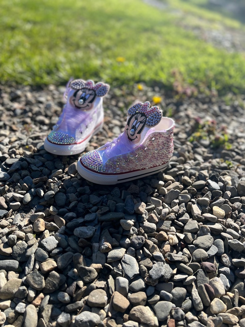 May include: A pair of custom high-top sneakers with a Minnie Mouse theme. The shoes are decorated with pink and silver glitter, rhinestones, and a Minnie Mouse face on the tongue. The shoes are sitting on a bed of small rocks.