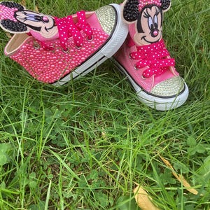 Puede incluir: Zapatillas rosas con adornos de Minnie Mouse. Los zapatos est&aacute;n cubiertos de peque&ntilde;as cuentas rosas y detalles de pedrer&iacute;a dorada en las punteras. Los cordones son de color rosa brillante con lunares blancos. Los zapatos est&aacute;n sobre hierba verde.