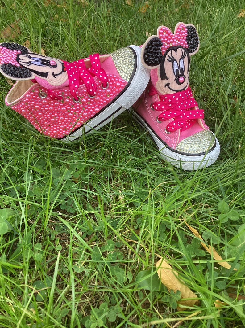 Puede incluir: Zapatillas rosas con adornos de Minnie Mouse. Los zapatos est&aacute;n cubiertos de peque&ntilde;as cuentas rosas y detalles de pedrer&iacute;a dorada en las punteras. Los cordones son de color rosa brillante con lunares blancos. Los zapatos est&aacute;n sobre hierba verde.