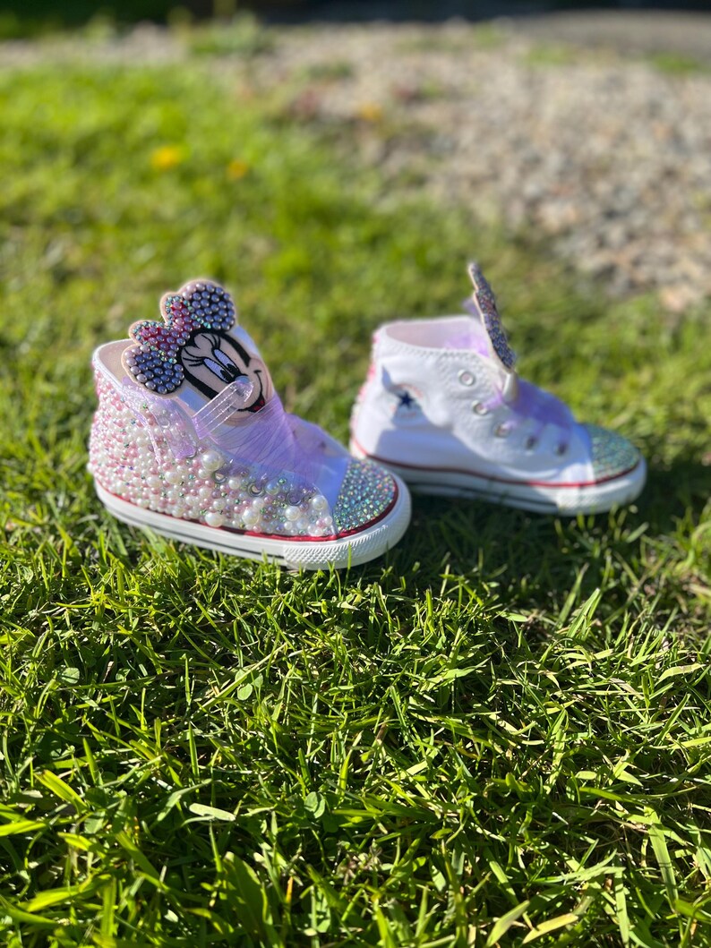 May include: A pair of custom-designed high-top sneakers, one featuring a Minnie Mouse design with pink and purple embellishments, the other with a star design. Both shoes have white uppers, red soles, and are decorated with rhinestones and pearls.