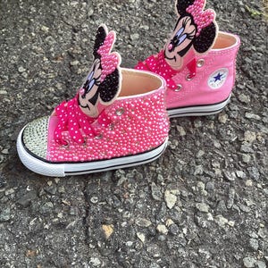 Puede incluir: Un par de zapatillas altas rosas con un dise&ntilde;o de Minnie Mouse. Las zapatillas est&aacute;n adornadas con pedrer&iacute;a rosa y tienen una suela blanca con una raya negra. La leng&uuml;eta del zapato tiene una cara y un lazo de Minnie Mouse.