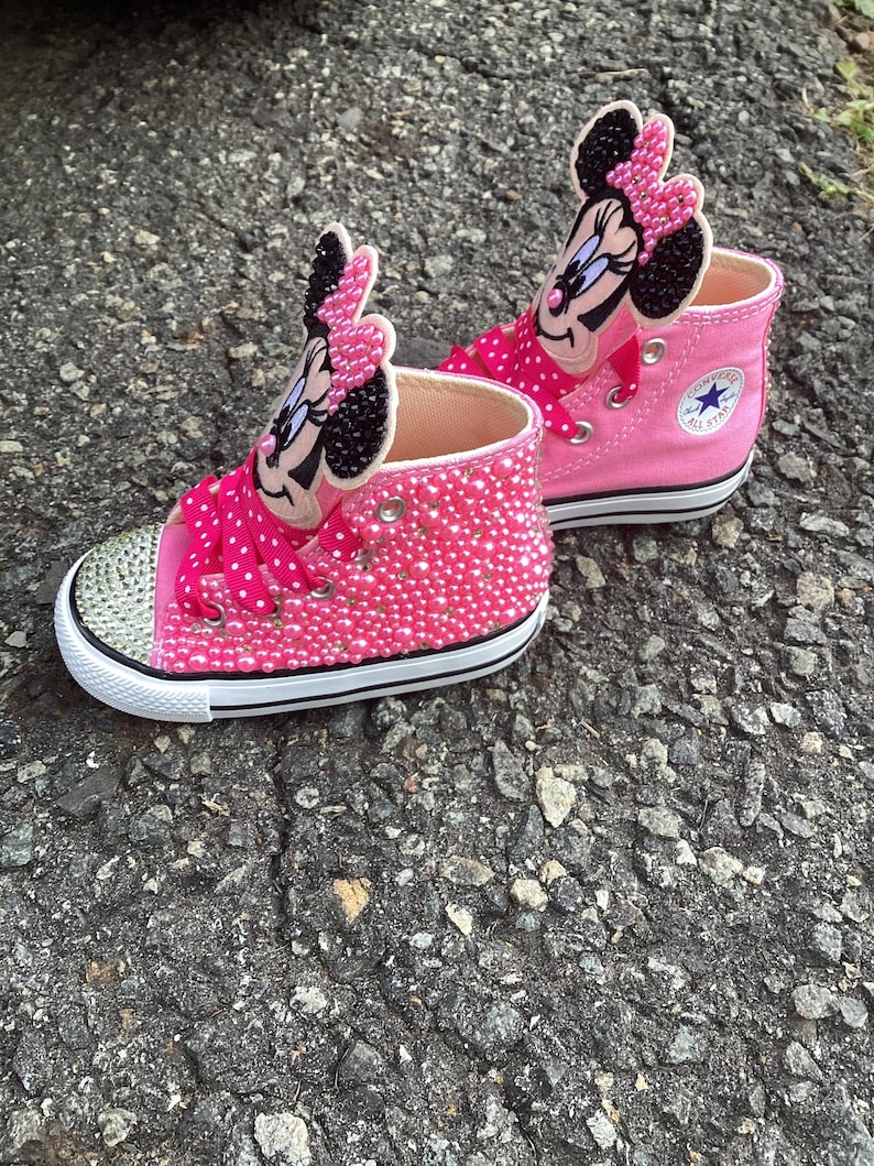 Puede incluir: Un par de zapatillas altas rosas con un dise&ntilde;o de Minnie Mouse. Las zapatillas est&aacute;n adornadas con pedrer&iacute;a rosa y tienen una suela blanca con una raya negra. La leng&uuml;eta del zapato tiene una cara y un lazo de Minnie Mouse.